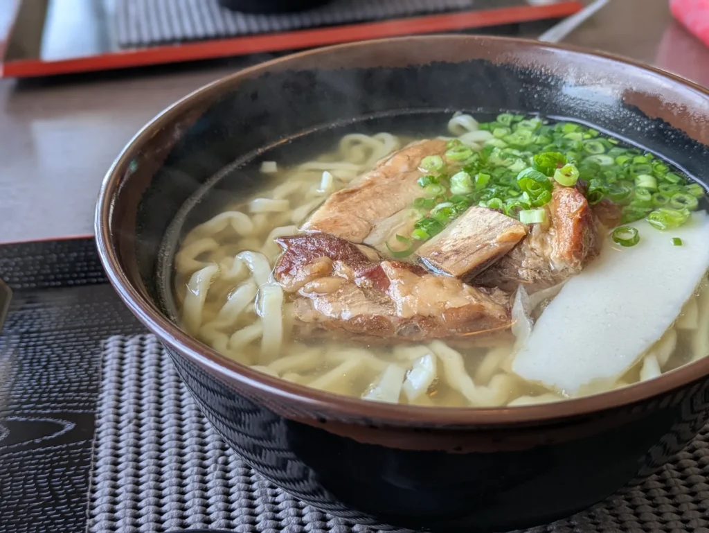 器に盛られた沖縄そば。透明な出汁に平打ち麺、三枚肉、かまぼこ、青ネギが確認できる。