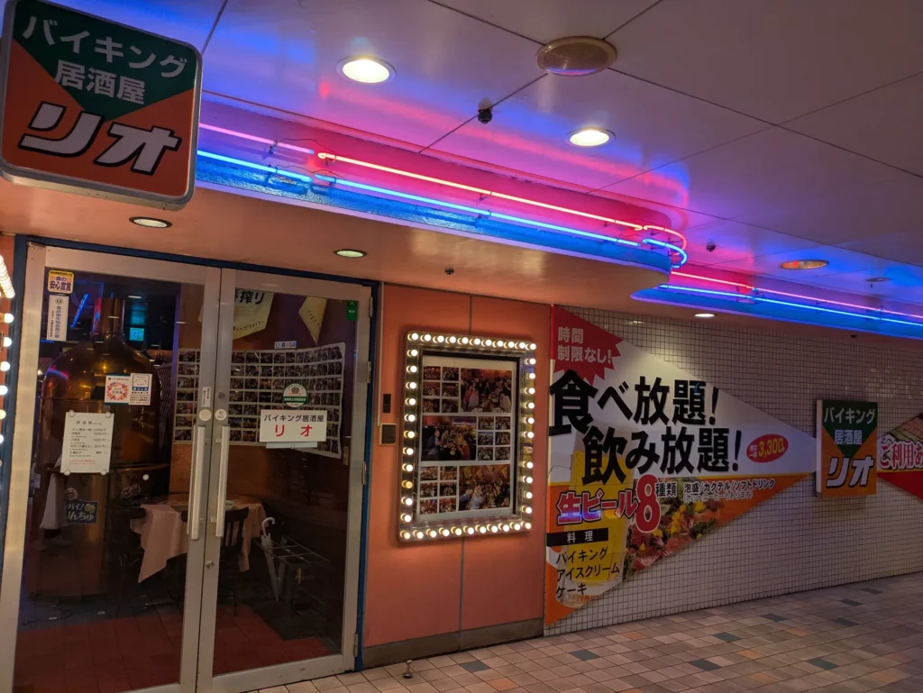 ネオンが輝く「バイキング居酒屋 リオ」の店舗入り口と、食べ飲み放題の料金が書かれた大きな看板